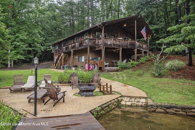 rear view of property featuring a lawn, a deck with water view, a patio, and a fire pit