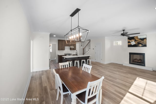 dining space with a fireplace with raised hearth, baseboards, light wood-type flooring, stairs, and a ceiling fan