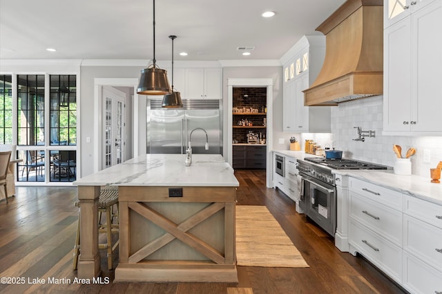 kitchen featuring custom exhaust hood, high end appliances, a center island with sink, white cabinets, and light stone counters