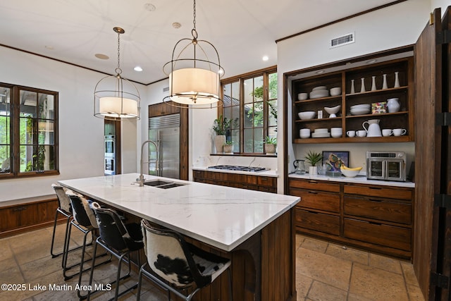 kitchen featuring built in refrigerator, a sink, stone tile flooring, a center island with sink, and decorative light fixtures