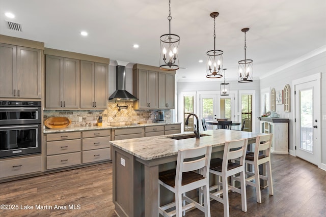 kitchen with black appliances, hanging light fixtures, a center island with sink, and wall chimney range hood