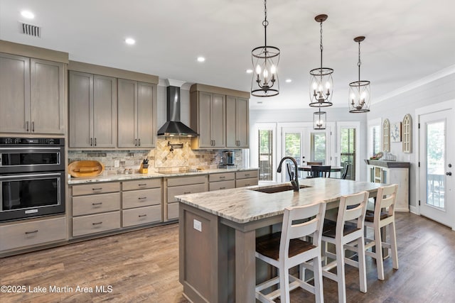 kitchen with wall chimney exhaust hood, a kitchen bar, light stone counters, hanging light fixtures, and a kitchen island with sink