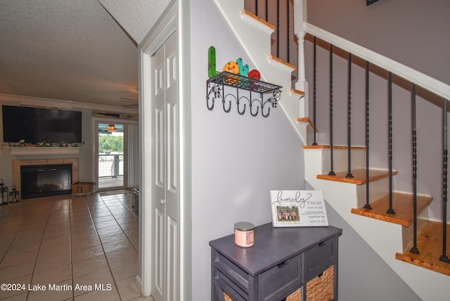 staircase featuring ornamental molding, a textured ceiling, ceiling fan, tile patterned flooring, and a tiled fireplace