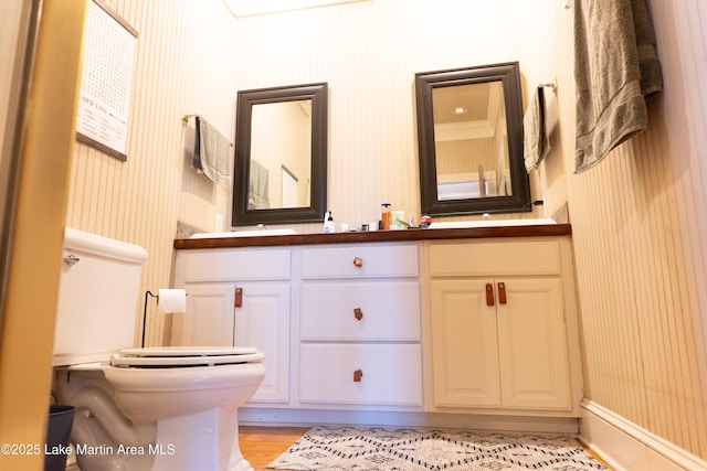 bathroom featuring double vanity, wallpapered walls, toilet, and wood finished floors