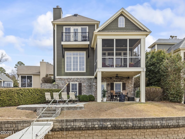 back of house featuring a balcony, a sunroom, stone siding, a chimney, and a patio area