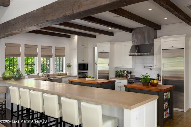 kitchen featuring kitchen peninsula, wall chimney range hood, built in appliances, beamed ceiling, and white cabinetry