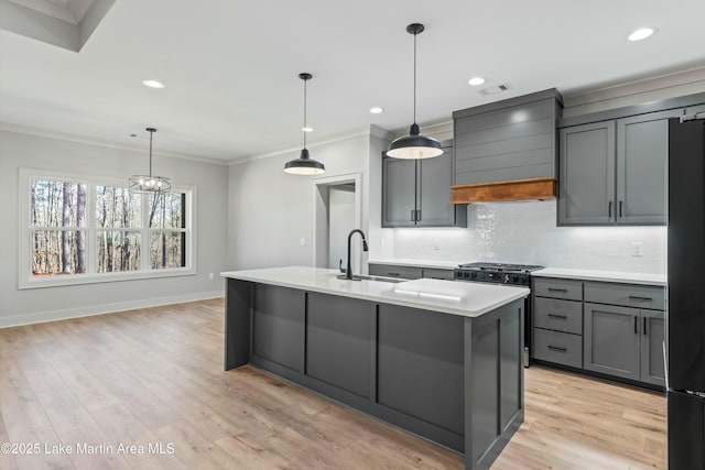 kitchen with a center island with sink, custom range hood, light countertops, pendant lighting, and a sink