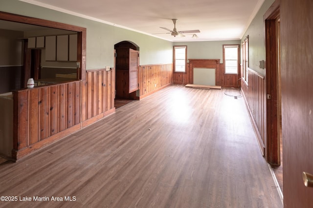 unfurnished living room with a wainscoted wall, arched walkways, ornamental molding, wood finished floors, and a ceiling fan