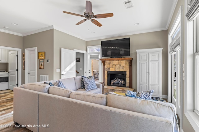 living area with wood finished floors, ornamental molding, a stone fireplace, visible vents, and a ceiling fan