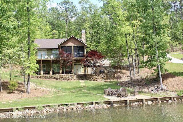 back of property featuring a sunroom, an outdoor fire pit, a deck with water view, a lawn, and a chimney