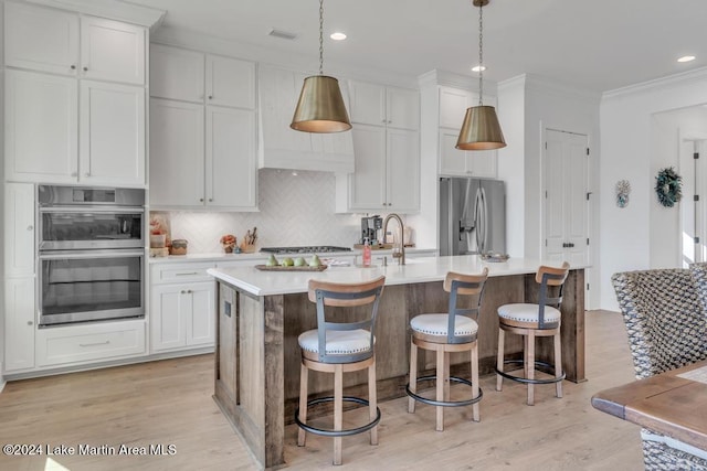 kitchen with light wood-type flooring, stainless steel appliances, a kitchen island with sink, pendant lighting, and white cabinets