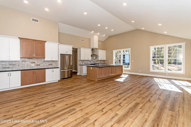 kitchen with tasteful backsplash, wall chimney exhaust hood, a large island with sink, white cabinetry, and stainless steel refrigerator