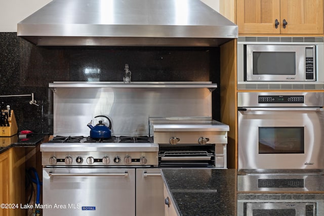 kitchen featuring decorative backsplash, appliances with stainless steel finishes, dark stone counters, wall chimney range hood, and light brown cabinets