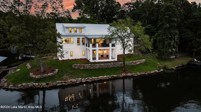 back house at dusk with a lawn, a balcony, and a water view