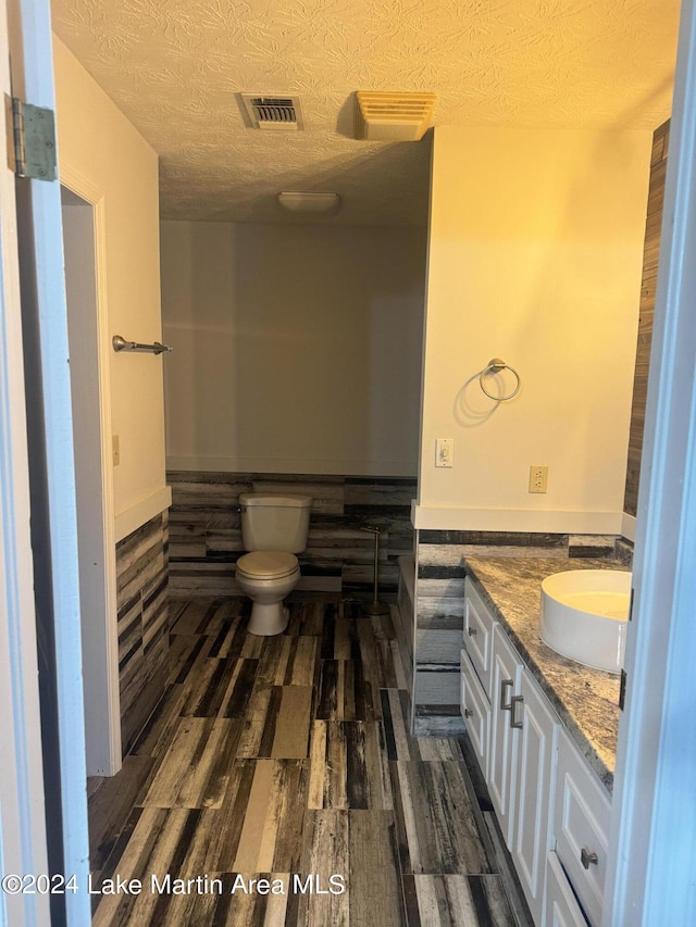 half bath with visible vents, toilet, wood finished floors, a textured ceiling, and vanity