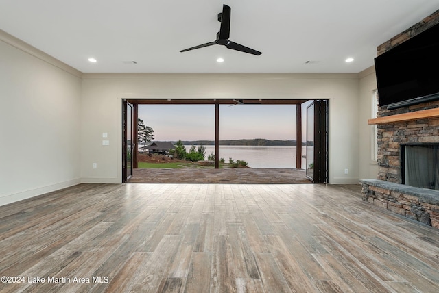 unfurnished living room featuring wood-type flooring, ceiling fan, a stone fireplace, and crown molding