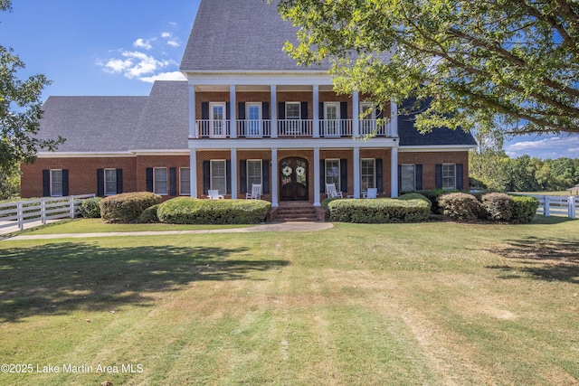 greek revival house featuring a balcony, roof with shingles, fence, a front lawn, and brick siding