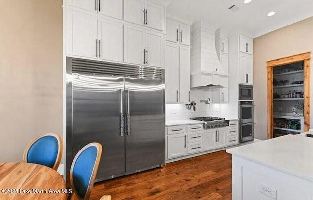 kitchen with white cabinets, decorative backsplash, custom exhaust hood, built in appliances, and dark wood-type flooring