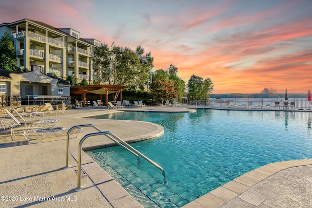 pool at dusk with a patio area, fence, and a community pool