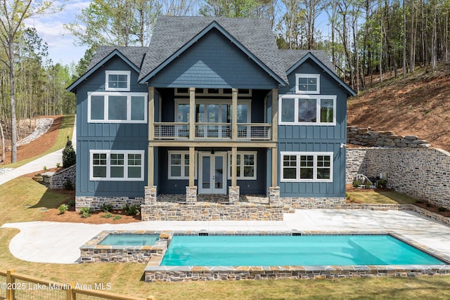 back of property featuring french doors, board and batten siding, a balcony, a pool with connected hot tub, and a shingled roof