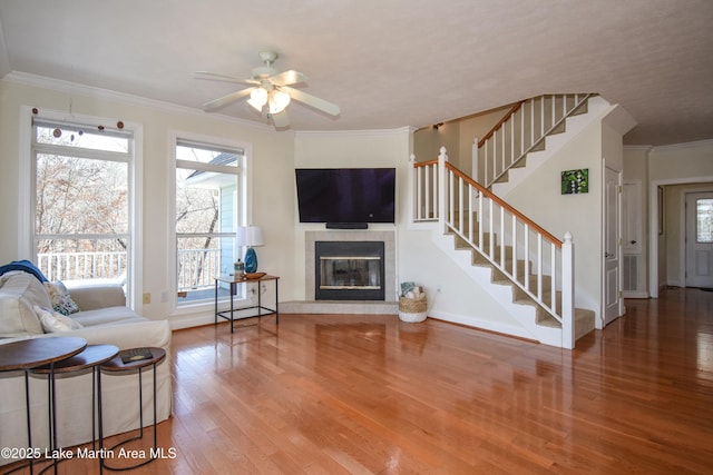 living area with stairway, wood finished floors, ceiling fan, a tile fireplace, and crown molding