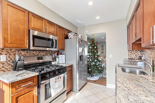 kitchen with brown cabinetry, appliances with stainless steel finishes, light stone countertops, a sink, and light tile patterned flooring