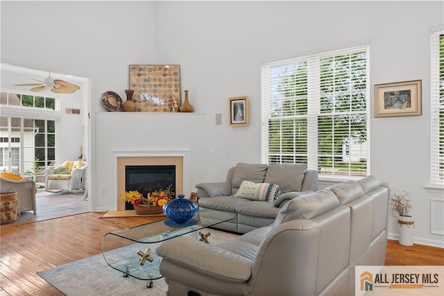 living room with wood finished floors, a ceiling fan, a glass covered fireplace, baseboards, and a towering ceiling