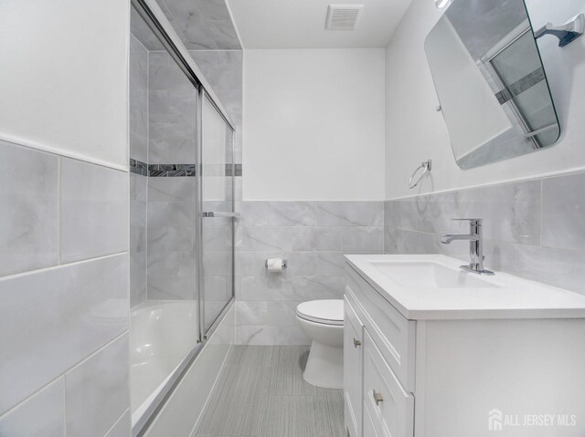 full bathroom with combined bath / shower with glass door, tile walls, tile patterned flooring, vanity, and toilet