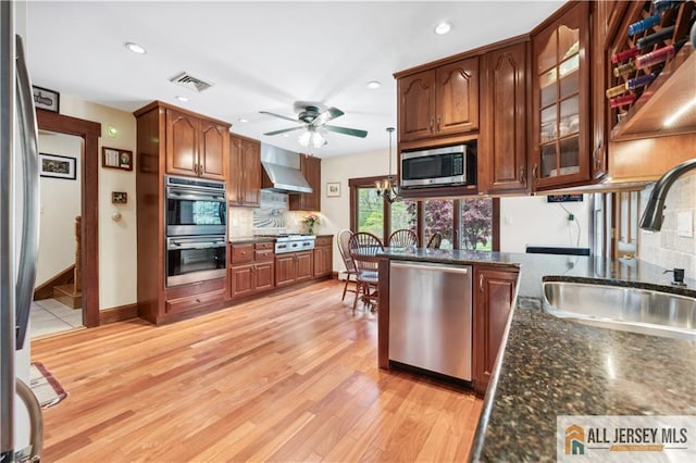 kitchen with stainless steel appliances, a sink, wall chimney range hood, ceiling fan, and tasteful backsplash