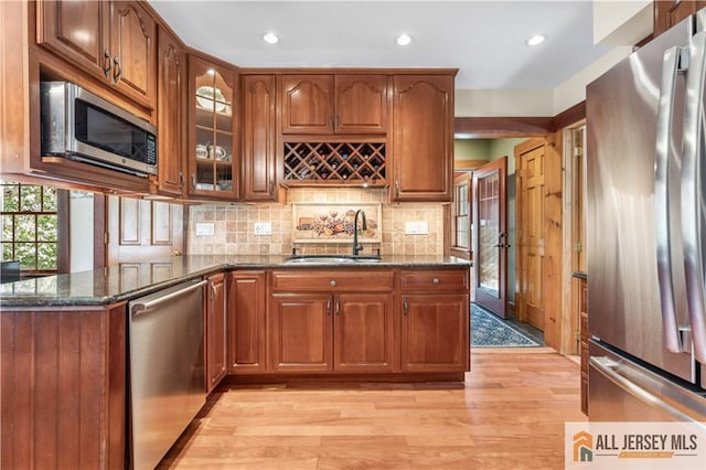 kitchen featuring appliances with stainless steel finishes, a sink, dark stone countertops, light wood-type flooring, and glass insert cabinets