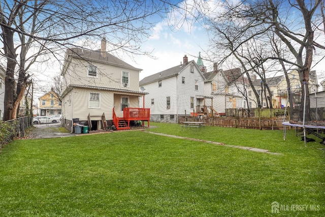 back of property with a deck, a trampoline, a fenced backyard, and a lawn