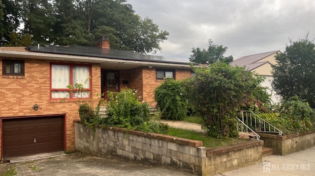 ranch-style home with an attached garage, solar panels, brick siding, driveway, and a chimney