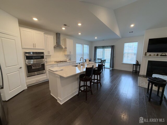 kitchen featuring wall chimney exhaust hood, a breakfast bar, white cabinetry, appliances with stainless steel finishes, and an island with sink