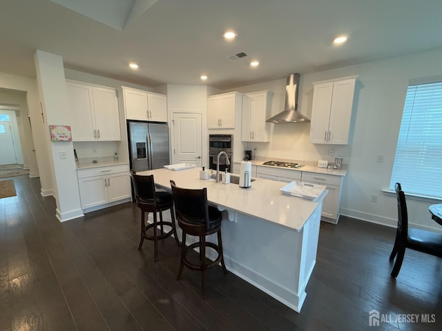 kitchen with white cabinetry, a center island with sink, dark wood-type flooring, appliances with stainless steel finishes, and wall chimney range hood