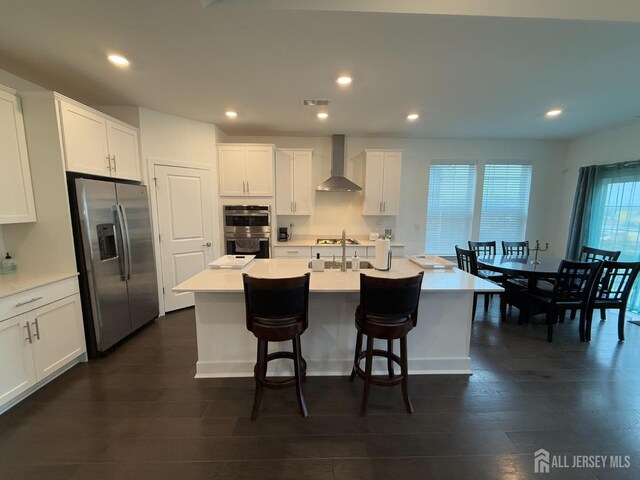 kitchen with white cabinetry, dark hardwood / wood-style flooring, an island with sink, stainless steel appliances, and wall chimney range hood