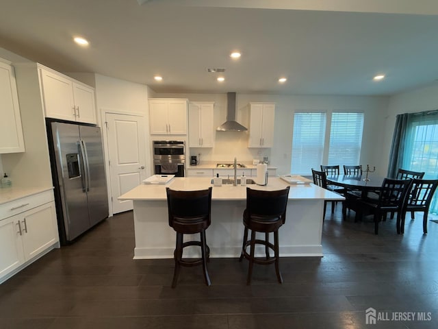 kitchen featuring appliances with stainless steel finishes, dark wood-type flooring, white cabinets, wall chimney range hood, and a kitchen island with sink