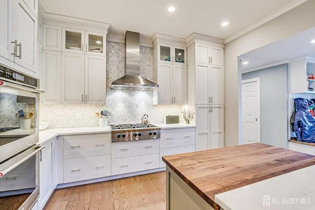 kitchen with wall chimney range hood, butcher block countertops, light wood-type flooring, ornamental molding, and stainless steel appliances