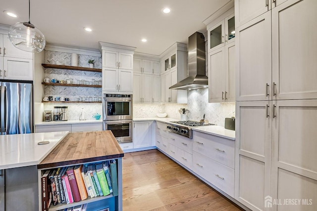 kitchen with butcher block countertops, light wood-style flooring, wall chimney exhaust hood, appliances with stainless steel finishes, and open shelves