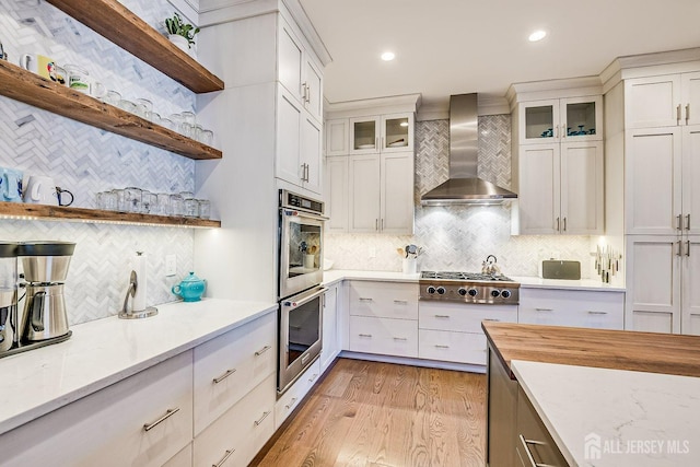 kitchen with open shelves, decorative backsplash, light wood-style floors, appliances with stainless steel finishes, and wall chimney exhaust hood