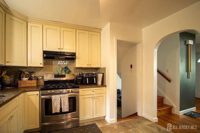 kitchen featuring stainless steel gas stove, under cabinet range hood, arched walkways, decorative backsplash, and baseboards