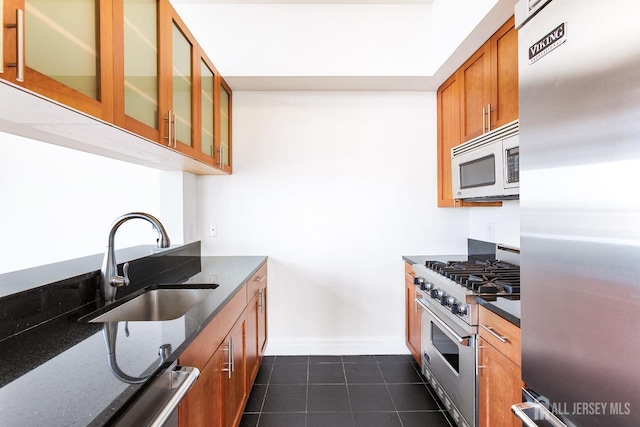 kitchen with stainless steel appliances, a sink, brown cabinets, dark stone countertops, and glass insert cabinets