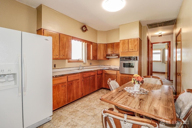 kitchen featuring oven, white refrigerator with ice dispenser, light countertops, brown cabinetry, and under cabinet range hood