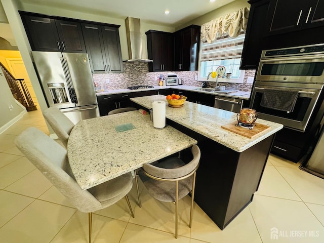 kitchen with light stone counters, wall chimney exhaust hood, dark cabinetry, a sink, and stainless steel appliances
