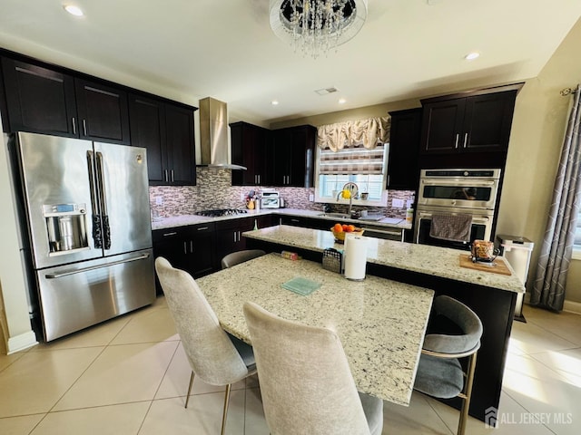 kitchen featuring wall chimney range hood, light stone countertops, dark cabinetry, stainless steel appliances, and a kitchen island