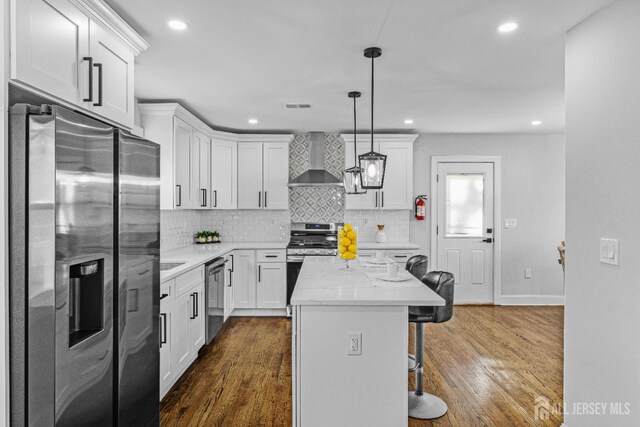 kitchen with appliances with stainless steel finishes, a center island, a kitchen bar, wall chimney range hood, and decorative light fixtures