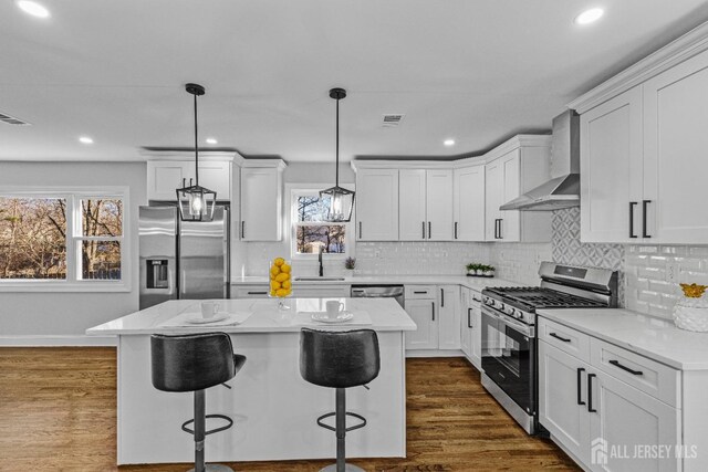 kitchen with wall chimney exhaust hood, decorative light fixtures, a kitchen island, white cabinetry, and appliances with stainless steel finishes