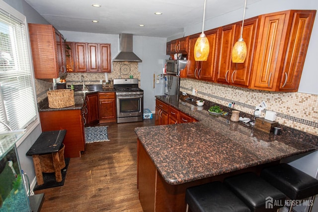 kitchen with pendant lighting, wall chimney range hood, stainless steel appliances, dark stone counters, and kitchen peninsula