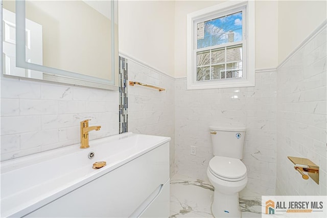 half bath featuring tile walls, toilet, marble finish floors, vanity, and a wainscoted wall