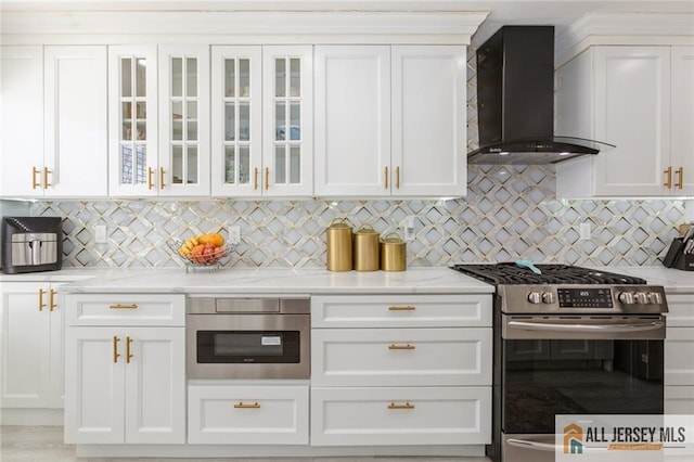 kitchen featuring white cabinetry, decorative backsplash, glass insert cabinets, appliances with stainless steel finishes, and wall chimney exhaust hood