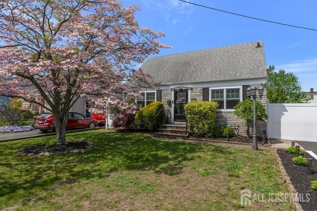 cape cod home featuring a shingled roof, fence, stone siding, and a front yard